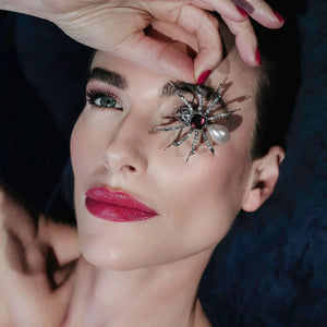 Model holding the Bill Skinner Pearl Spider Brooch over her eye, showcasing the brooch’s luxurious amethyst and pearl detailing against her glowing skin and bold red lips.