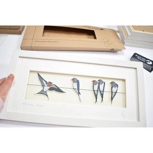 Hand Holding Framed ‘Welcome Home’ Print – A person holds the ‘Welcome Home’ framed artwork, showing the swallows perched on a wire with one flying in. The background includes packaging materials.