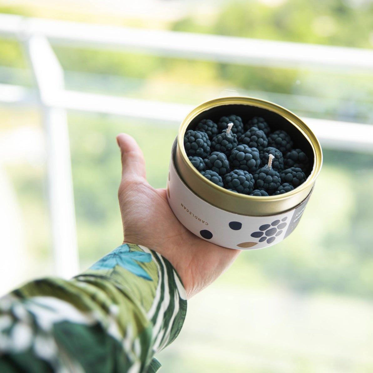 Hand holding sealed Blackberry CandleCan in natural daylight, showing size and playful design against blurred green background.