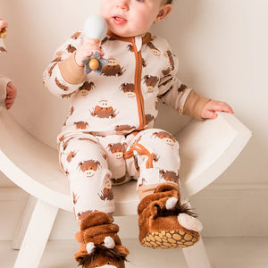 Baby sitting in a chair wearing the Neutral Hamish Highland Cow romper with matching cow slippers, looking up and smiling.