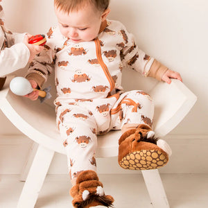 Baby in cow print romper and slippers seated in a modern white chair, gazing down at another baby’s toy.