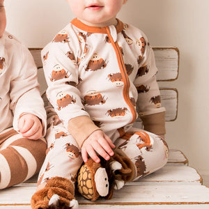 Baby sitting on a bench in the Neutral Hamish Highland Cow zip-up romper and slippers, facing forward with a gentle smile.