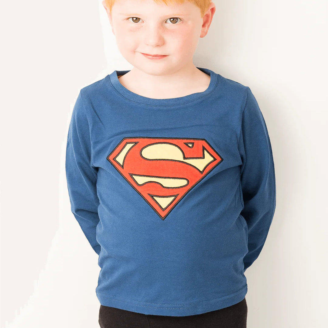 Little boy wearing Superman™ “Saves the Day” blue top with red and yellow ‘S’ shield, hands behind back, smiling.