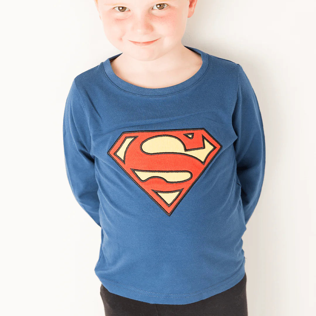Child wearing Superman™ blue long-sleeved top with iconic ‘S’ shield, smiling playfully.