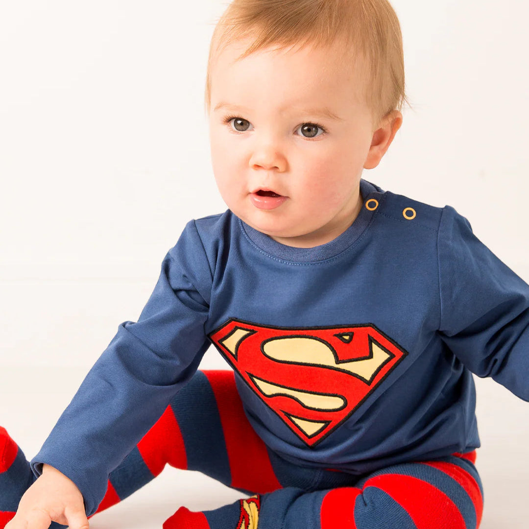 Toddler in Superman™ top with striped leggings, waving while seated on floor.