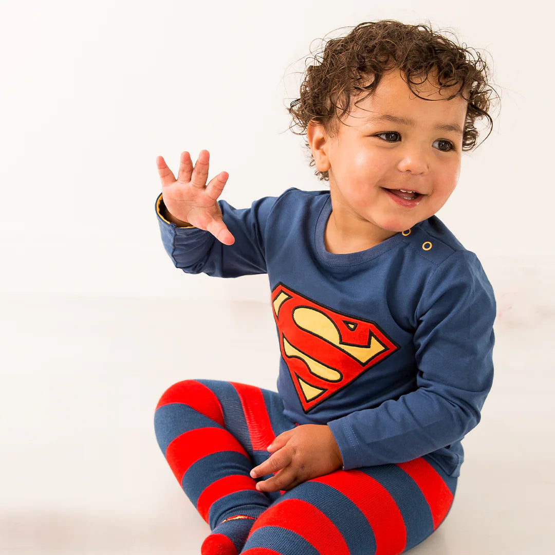 Flat lay of Superman™ blue long-sleeved top with yellow-trimmed neckline and bold applique ‘S’ shield, on red and grey background.
