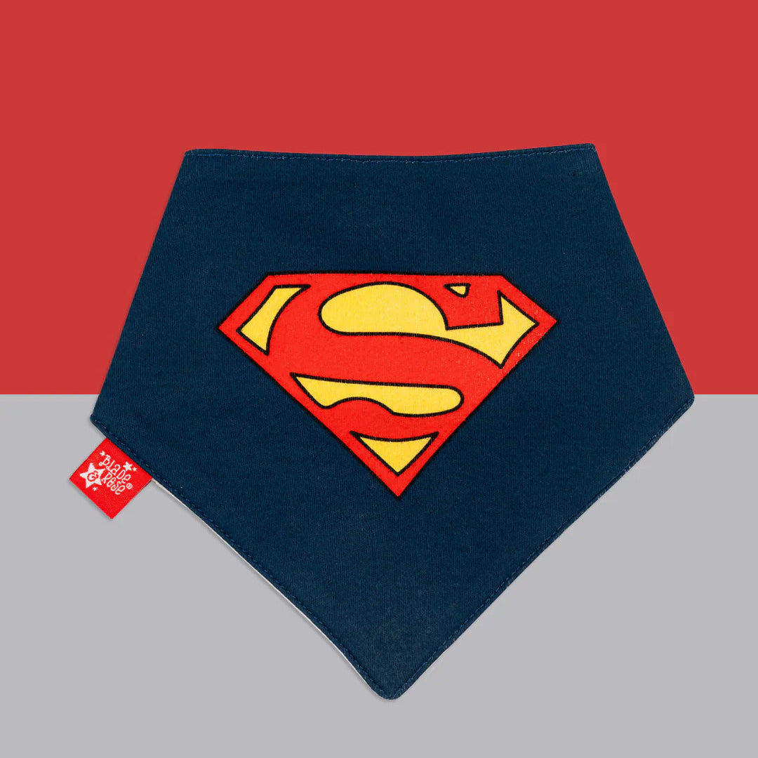 Navy Superman™ baby bib with large red and yellow “S” shield emblem, displayed flat on a red and grey background.