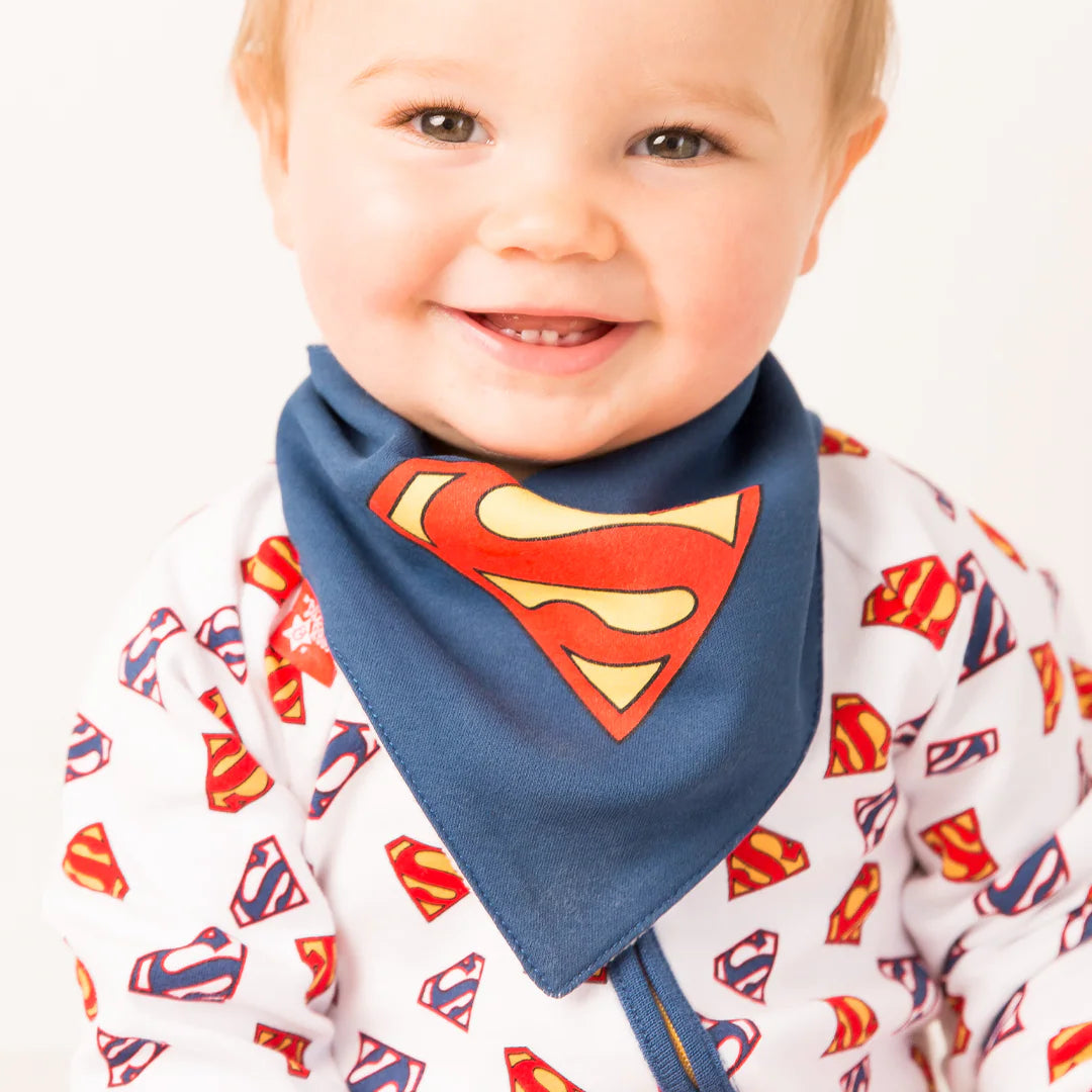 Smiling baby wearing navy Superman™ bib with large “S” shield logo, paired with a white Superman™ logo print outfit.