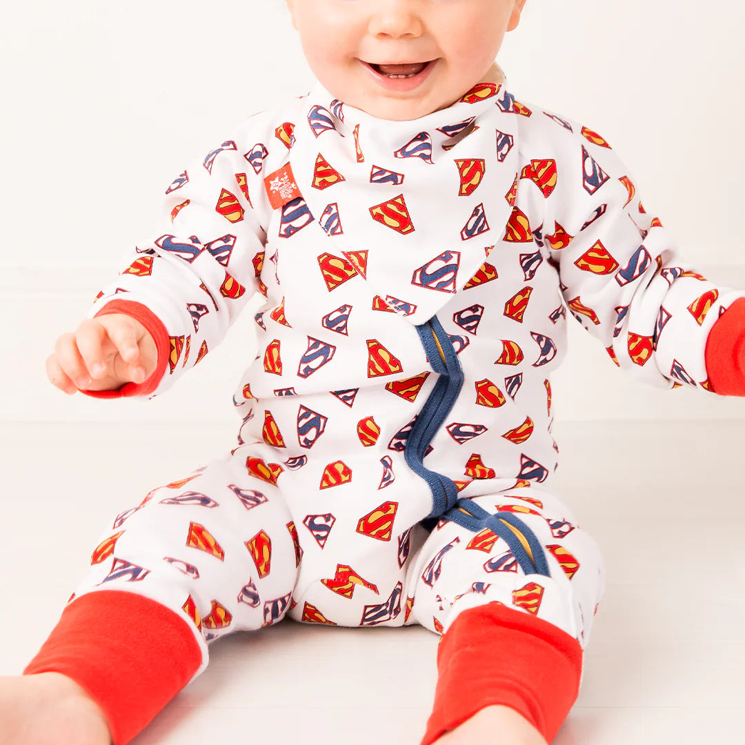 Smiling baby wearing Superman Save The Day Zip Up Romper with white cotton base, all-over red and yellow Superman shield print, bright red cuffs, and navy zip.