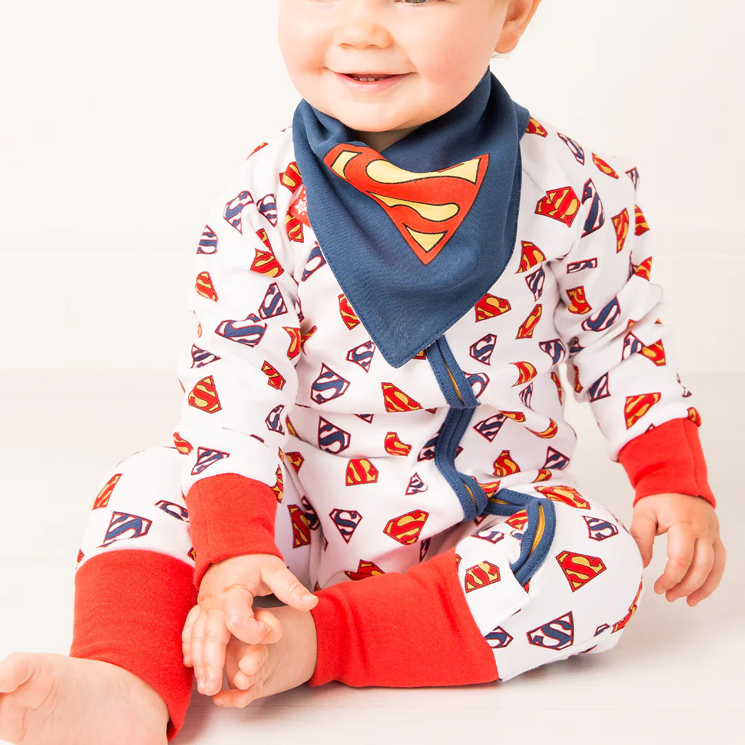 Baby wearing Superman Save The Day Zip Up Romper paired with navy bib featuring large Superman shield logo, white cotton fabric with all-over shield print.