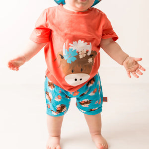 Baby standing in full Bonnie Highland Cow summer outfit with printed shorts and hat.