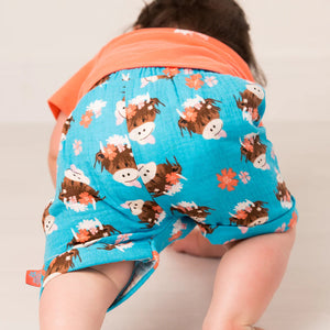 Baby wearing Bonnie Highland Cow muslin shorts crawling away in bright printed shorts.