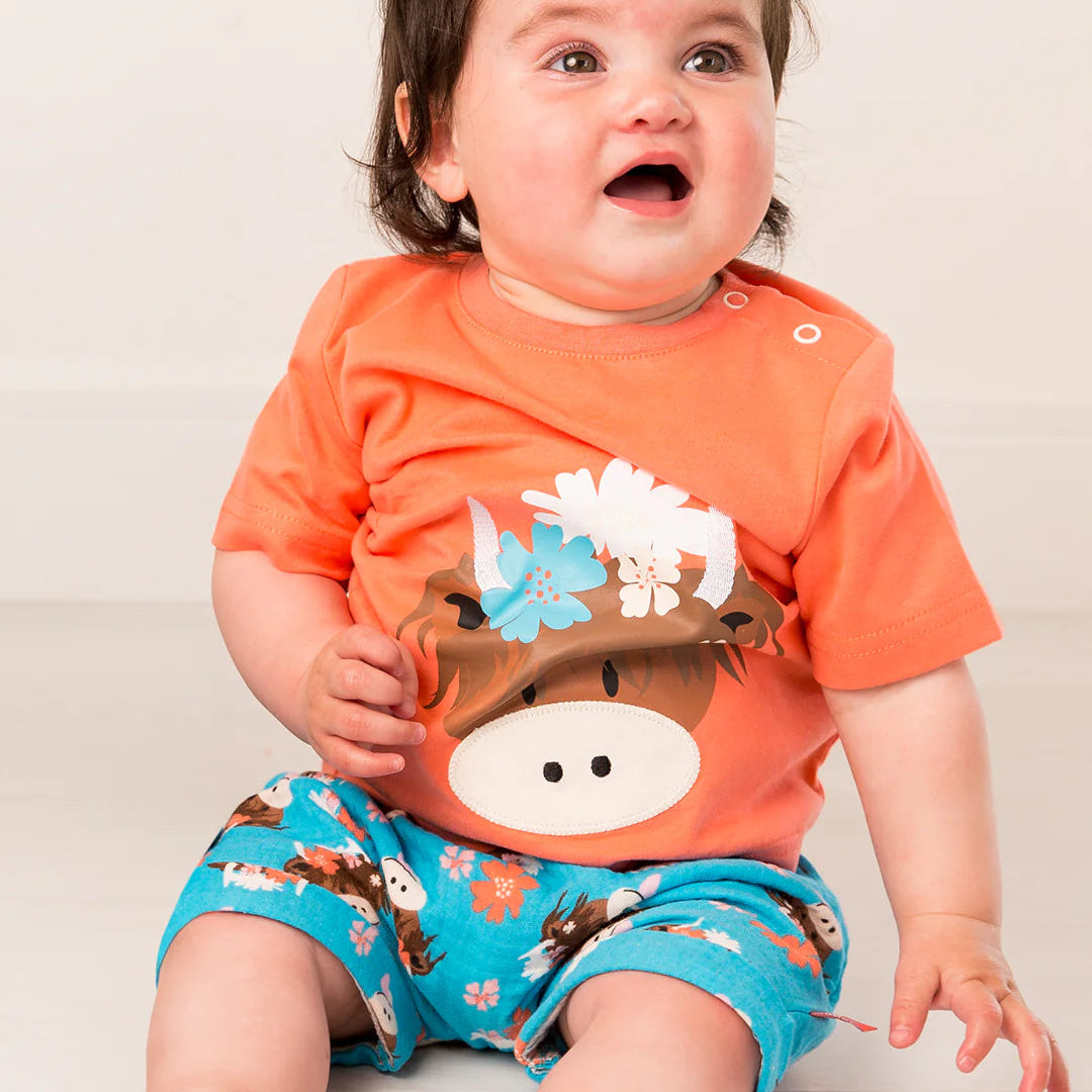 Baby girl wearing orange Bonnie Highland Cow t-shirt with turquoise floral cow print shorts, sitting on a light floor.