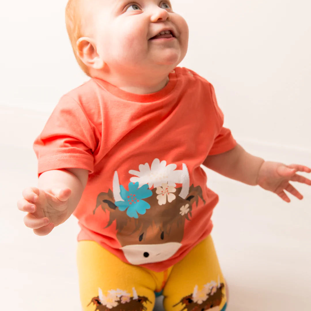 Smiling baby in Bonnie Highland Cow orange t-shirt paired with yellow cow print leggings, kneeling on a light background.