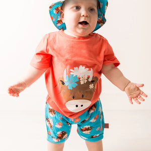 Toddler standing in orange Bonnie Highland Cow t-shirt and matching turquoise cow print shorts and bucket hat.