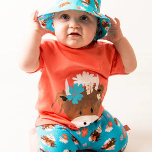 Baby in Bonnie Highland Cow orange tee and turquoise cow print shorts, adjusting a matching blue bucket hat.