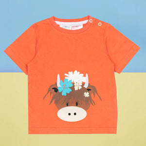 Flat lay of orange Bonnie Highland Cow baby t-shirt featuring floral cow face print, against a colourful backdrop.