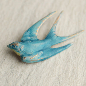 A handcrafted blue bird swallow brooch with a brass base, hand-enameled in cornflower blue with gold accents, lying on a light textured surface.