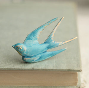 A vintage-style blue swallow brooch with gold detailing perched on the edge of a linen-covered book, showcasing its intricate enamel finish.