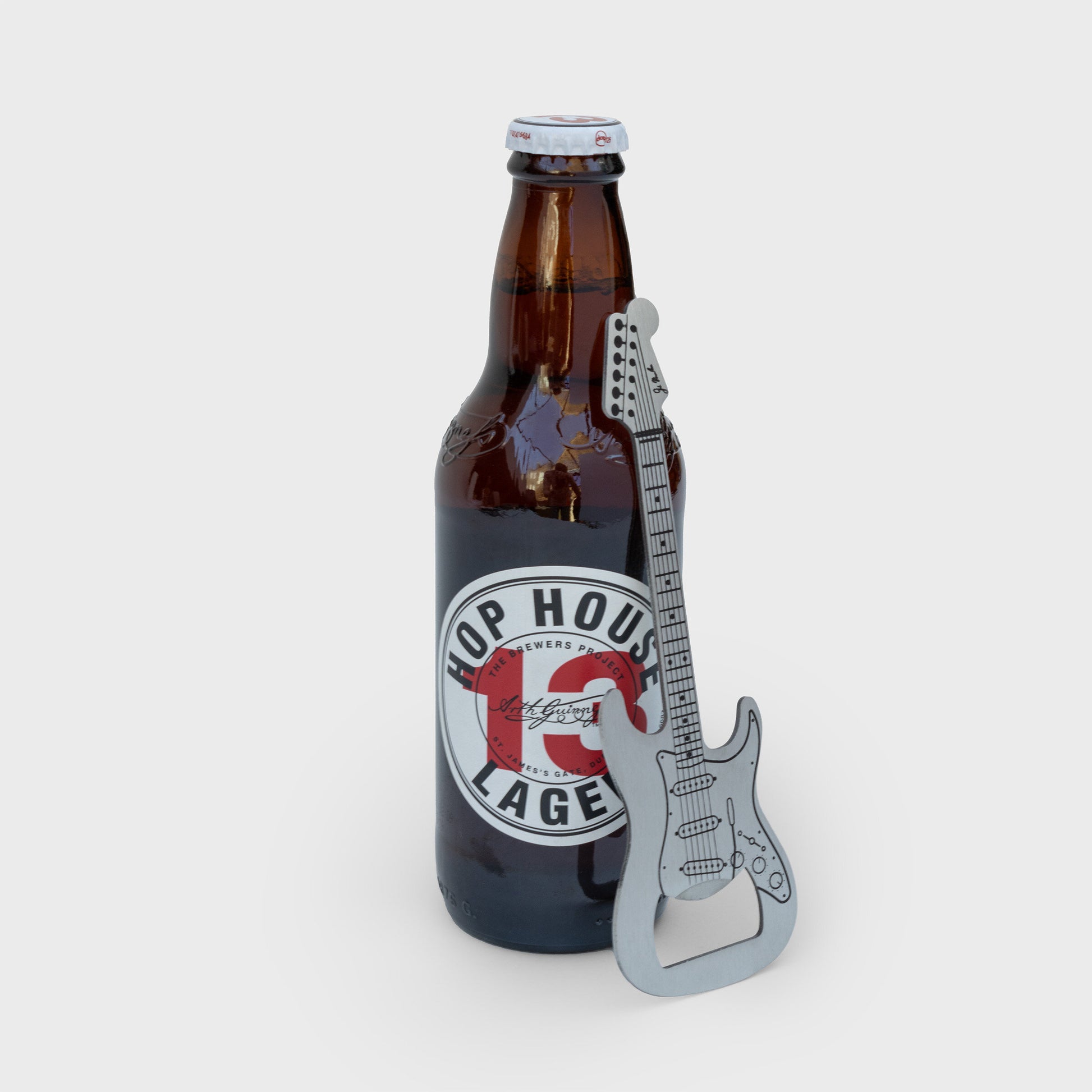Stainless steel guitar-shaped bottle opener propped against a Hop House 13 Lager bottle, with product detailing clearly visible.