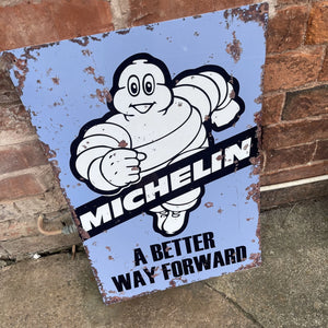 Vintage-style Michelin “A Better Way Forward” metal sign leaning against a red brick wall, featuring the iconic Michelin Man in black and white on a blue background.