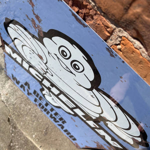 Close-up of the Michelin “A Better Way Forward” metal sign with a retro design, showing the Michelin Man running across a vintage blue surface with distressed edges.