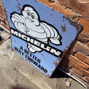 Angled view of the Michelin “A Better Way Forward” metal sign placed outdoors, showcasing its weathered finish and bold typography for a classic vintage look.