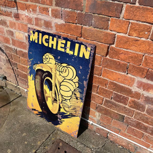 Vintage-style Michelin Motorcycle Tires metal sign leaning against a red brick wall, featuring the iconic Michelin Man riding a bike in bold yellow and navy tones.