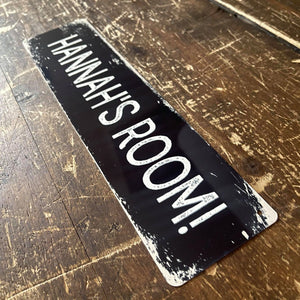 A custom metal sign displaying “HANNAH’S ROOM!” in white text on a black background, placed on a wooden surface, showing a unique personalised design.