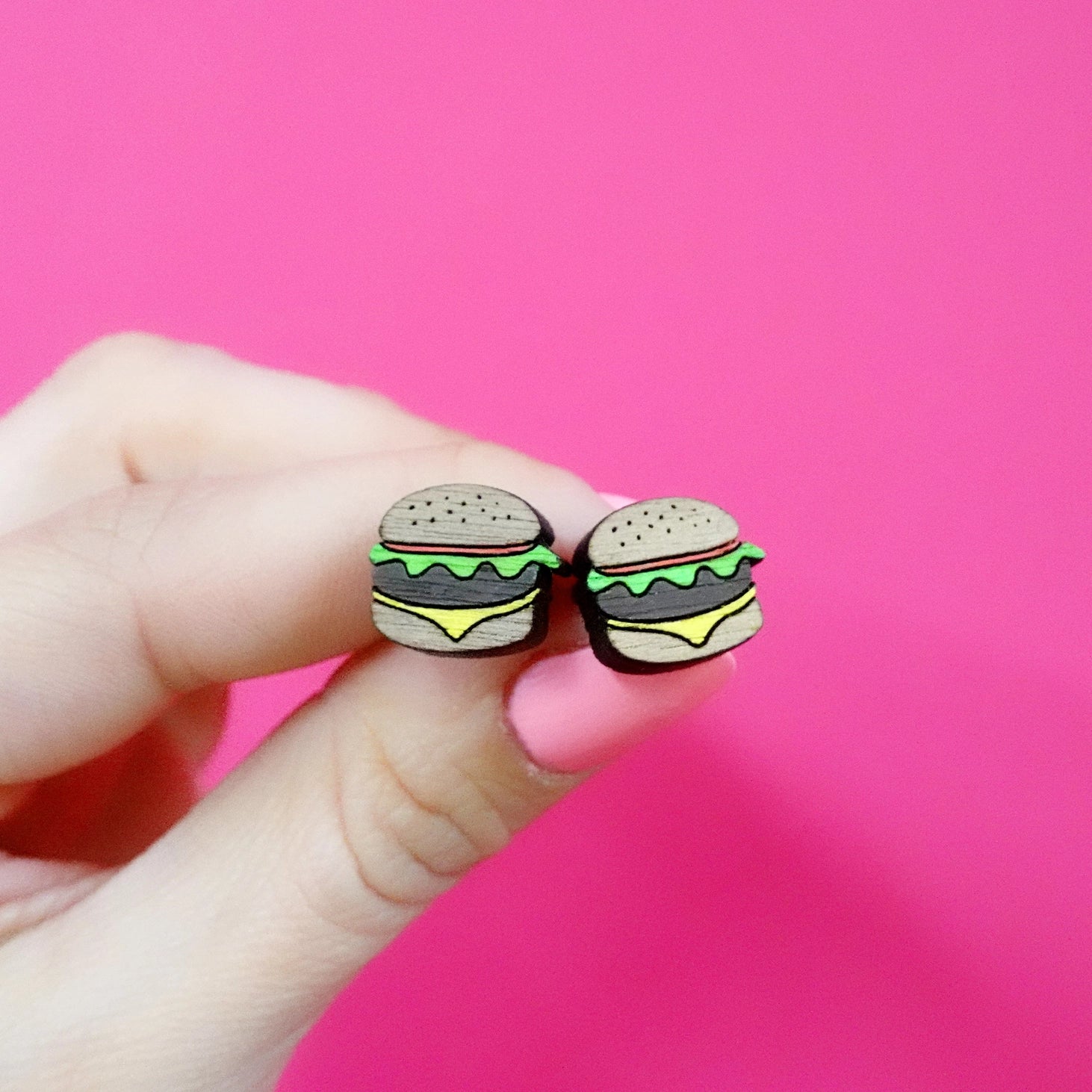 Close-up of hand holding a pair of handmade wooden burger stud earrings with detailed layers of bun, lettuce, cheese, and patty against a bright pink background.