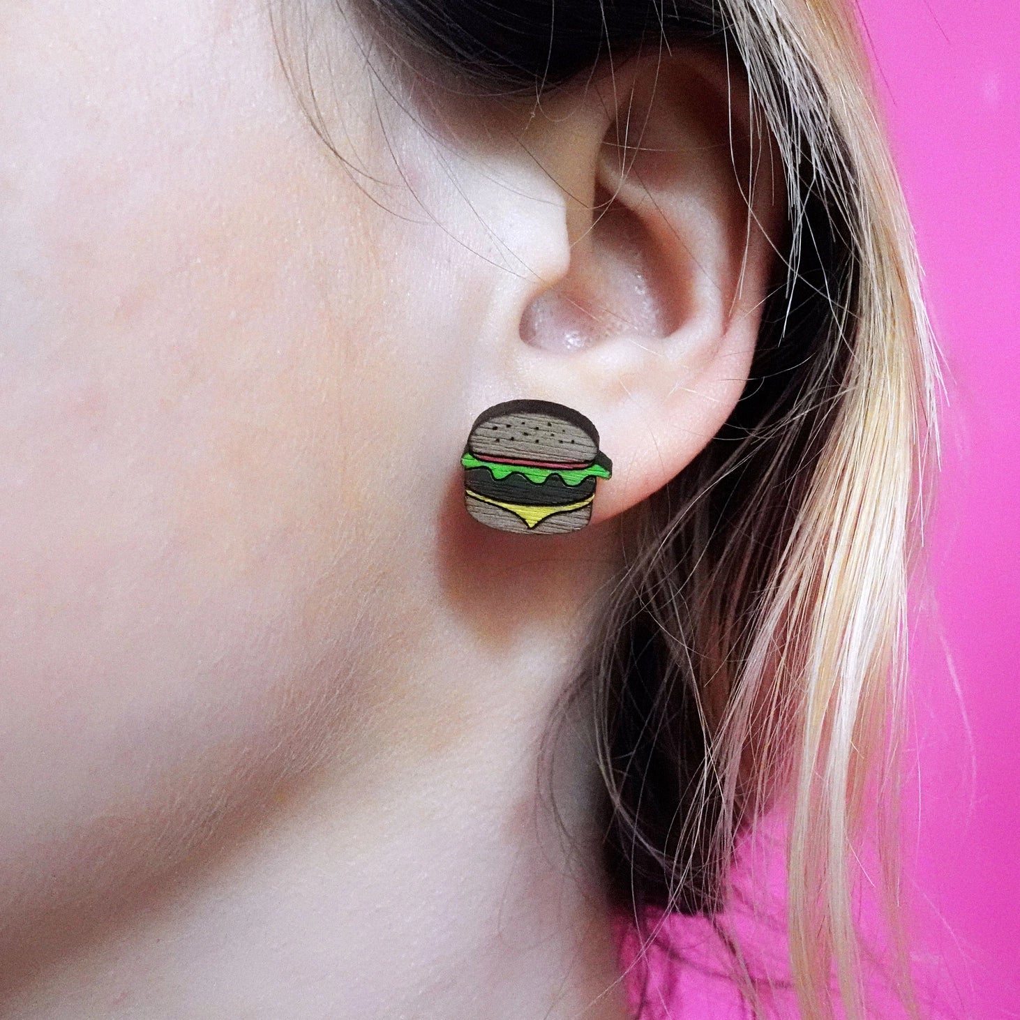 Burger-shaped wooden stud earring worn on ear, showcasing the quirky fast food design with green lettuce, yellow cheese, and engraved sesame seed bun.