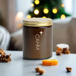 CAHM Clove, Orange & Cedarleaf candle styled on table with orange slice and spices, festive background bokeh.