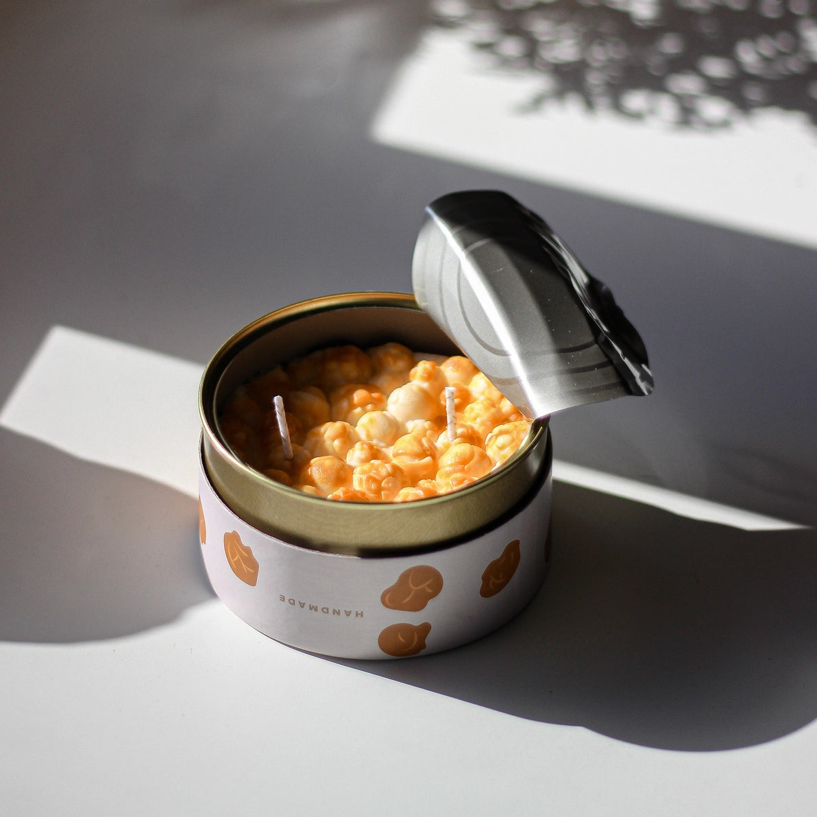 Caramel Popcorn candle with opened tin lid catching natural sunlight, highlighting lifelike popcorn detail and dual wick design.