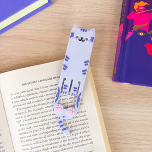 Flexible cat-shaped reading light clipped to an open book, showing its bookmark functionality and cute paw design on a wooden surface with books nearby.