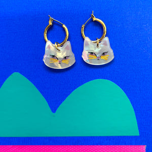 Catnip Cat Hoop Earrings on a blue background – A close-up of the acrylic cat face earrings on a bright blue backdrop with green and pink design elements.