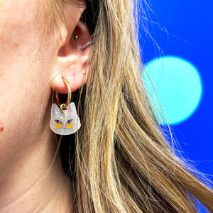 Close-up of a person wearing Catnip Cat Hoop Earrings – A stylish gold hoop earring featuring an acrylic cat face charm, worn on an ear with a vibrant blue background.