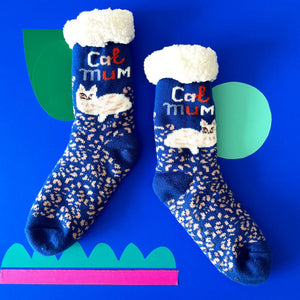 Flat lay of Catnip Cozy Slipper Socks on a blue background, showing bold “Cat Mum” lettering, white fluffy cuffs, and reclining white cat design with pink leopard-style pattern.