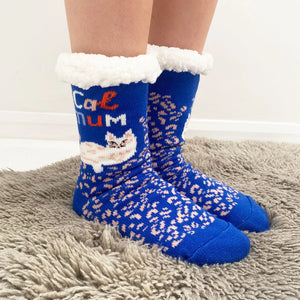 Person wearing Catnip Cozy Slipper Socks standing on a grey rug, showing off the soft white ankle cuffs, blue base, and “Cat Mum” slogan with cat motif.