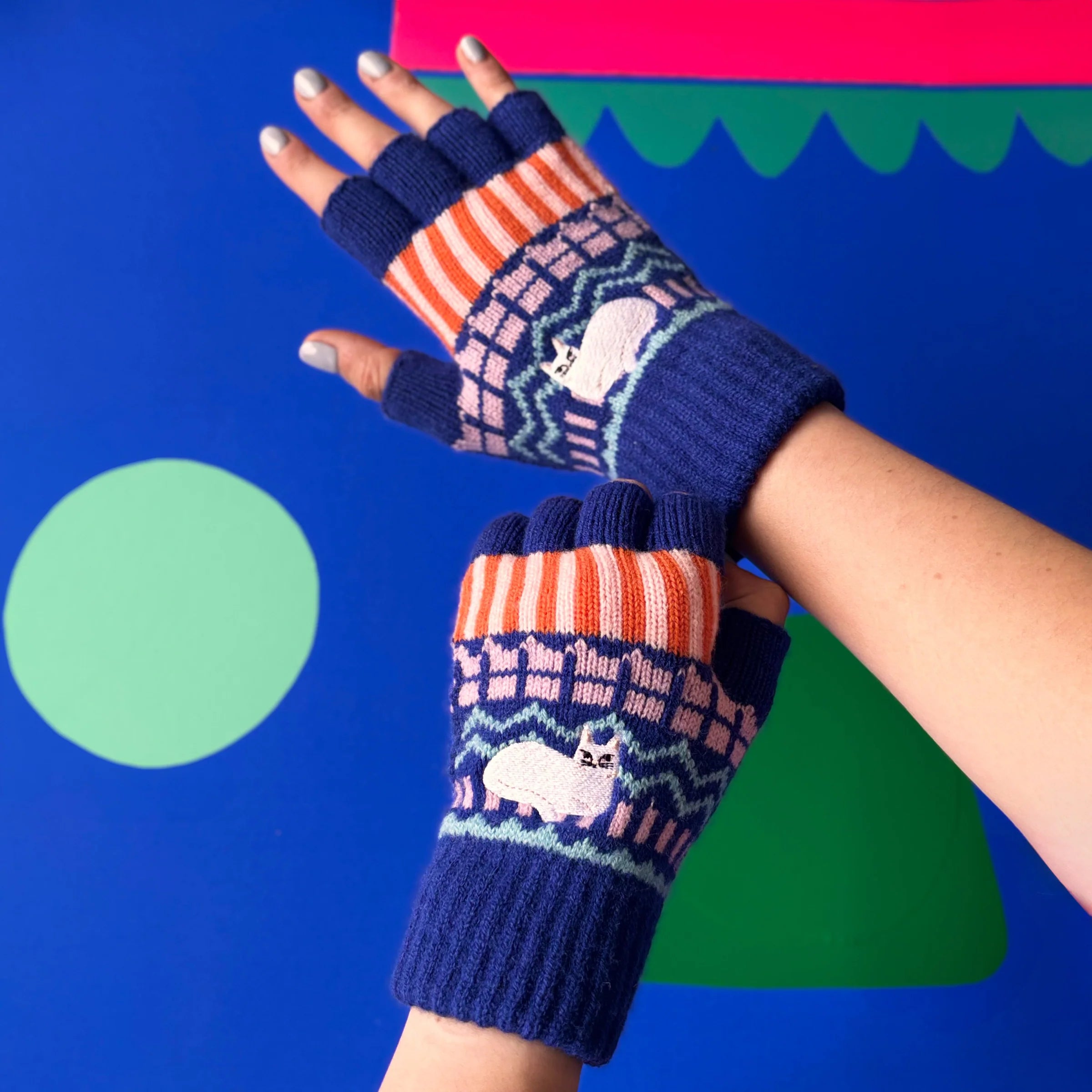 Colourful Catnip Fair Isle fingerless gloves with striped cuffs and embroidered cat motif, model wearing gloves against bright blue and pink backdrop.