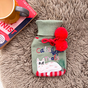 Green knitted Cats Before Twats hot water bottle with embroidered cat and red pom-poms on a fluffy rug.