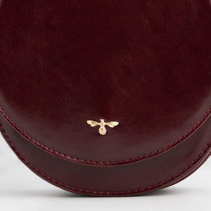 Detailed shot of gold bee emblem on front flap of Fable England Cherry Red Saddle Bag in rich cherry red tone.