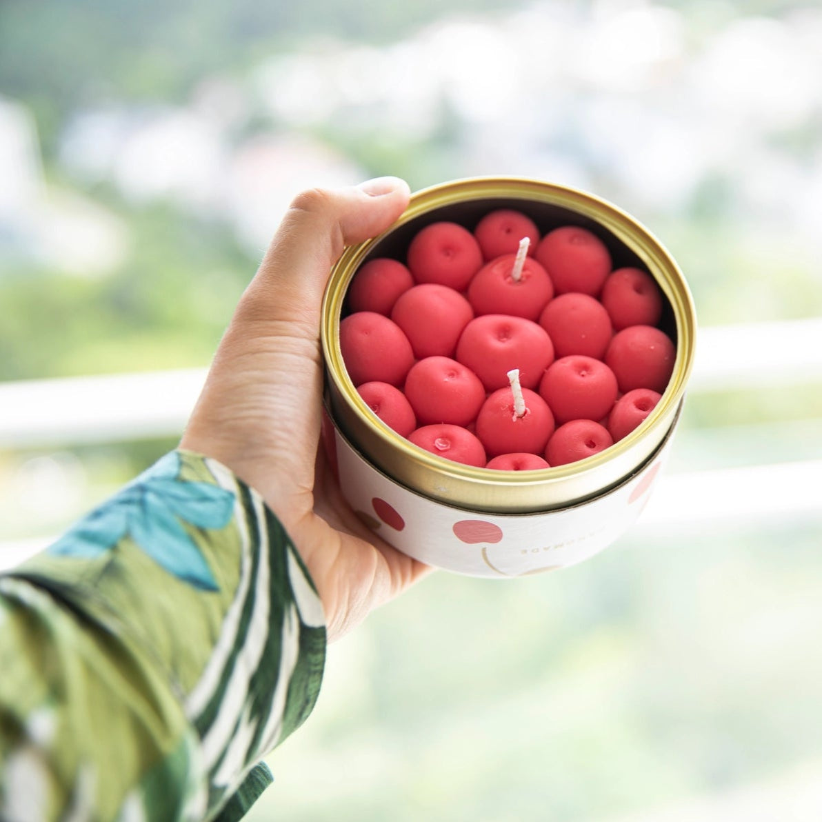 Hand holding open cherry CandleCan with vibrant cherry-shaped wax candles and scenic background.