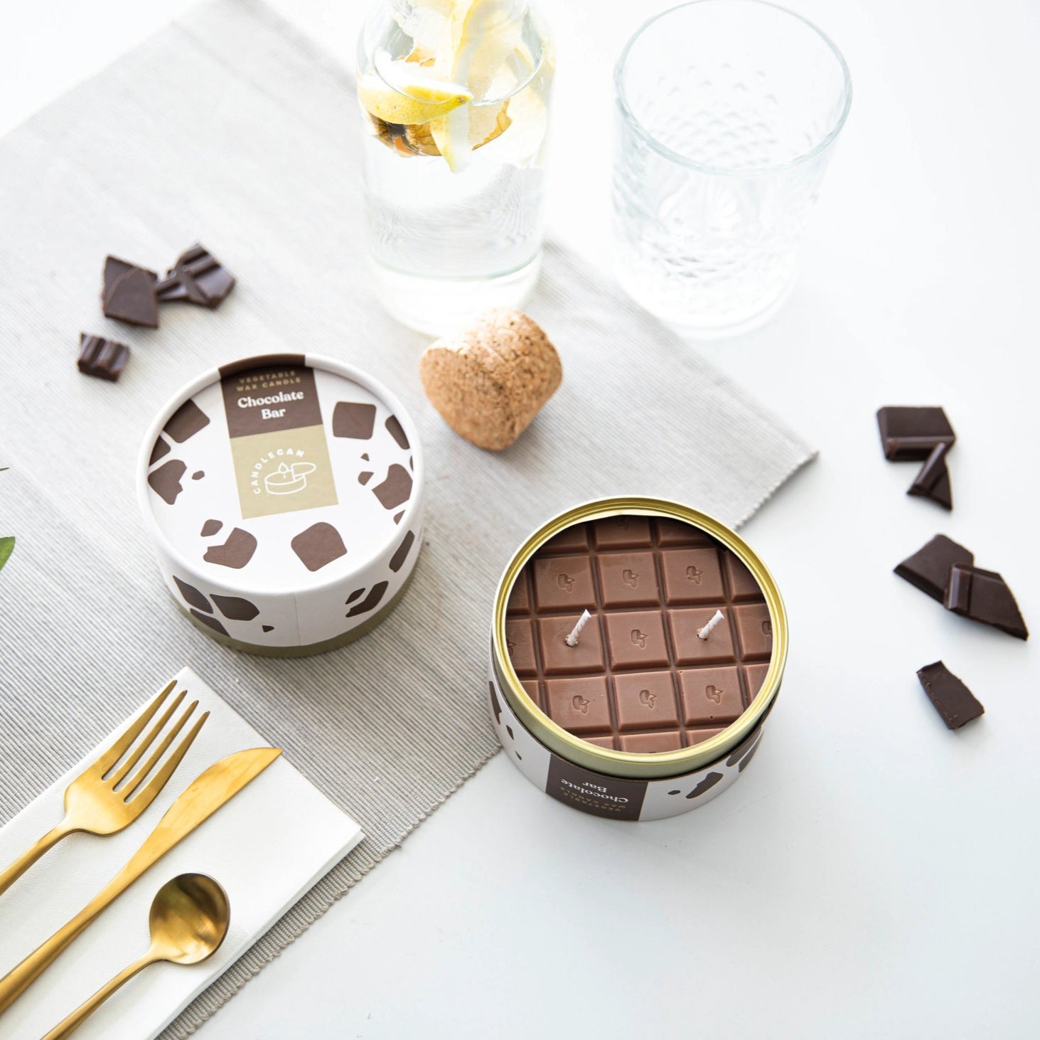 Flat lay of chocolate bar candle and tin beside a water carafe, cutlery, and chocolate pieces—styled for a dining table.