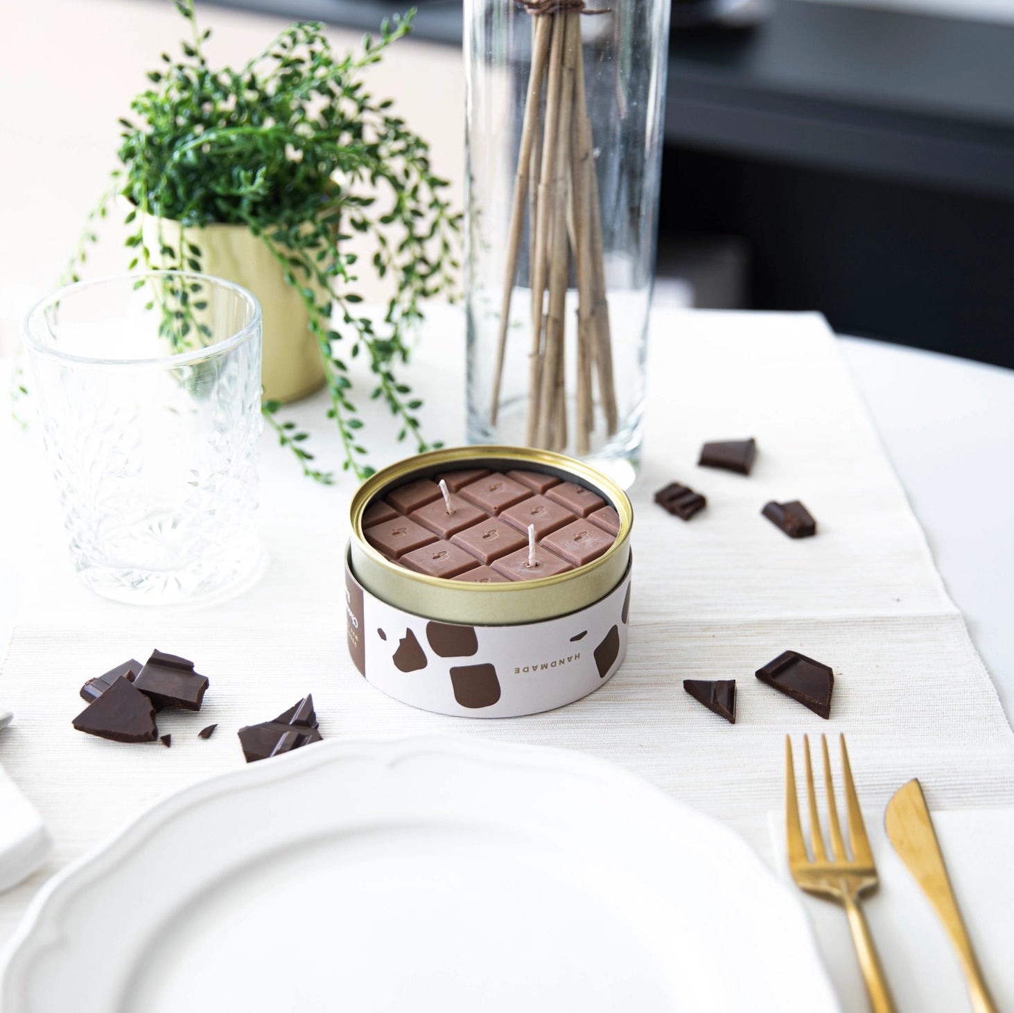 Chocolate scented candle on white tablecloth with gold cutlery and broken chocolate chunks, styled with modern home décor.