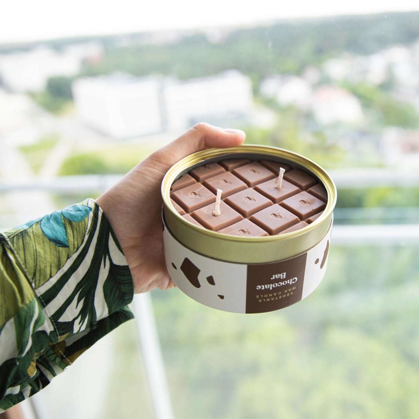 Hand holding open chocolate bar candle tin near window, highlighting realistic design and gourmet-style presentation.