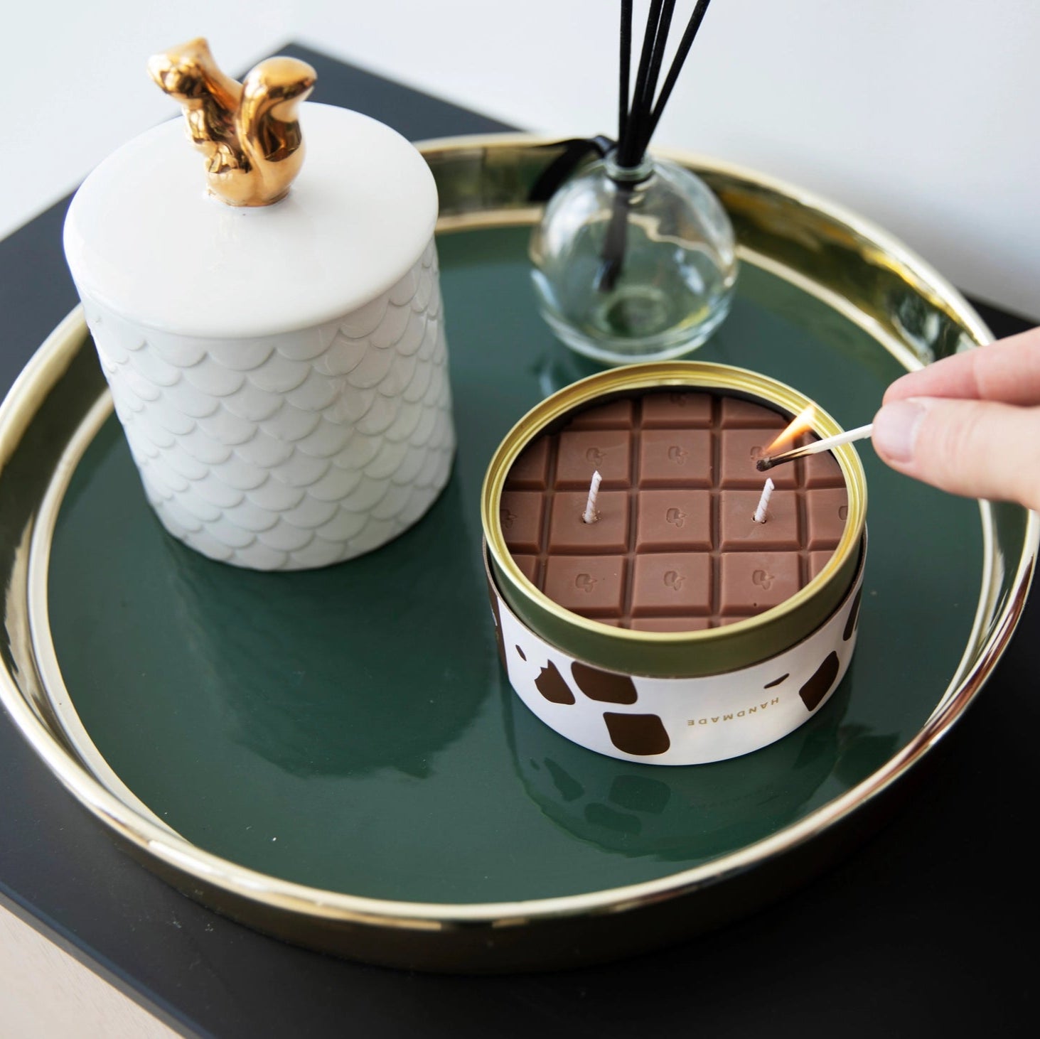 Chocolate bar candle being lit with a match, styled on a dark green tray with decorative homeware.