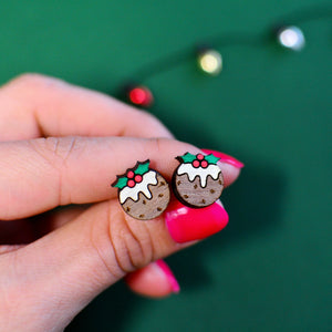 Close-up of hand holding wooden Christmas pudding stud earrings with holly detail, on a festive green background.