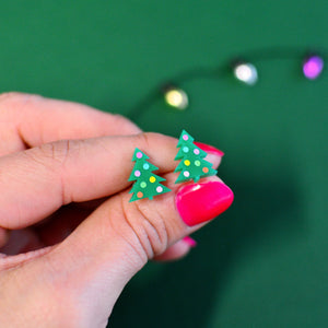 Person holding miniature Christmas tree stud earrings made from green Perspex acrylic with colourful hand-painted baubles, shown against a green background.