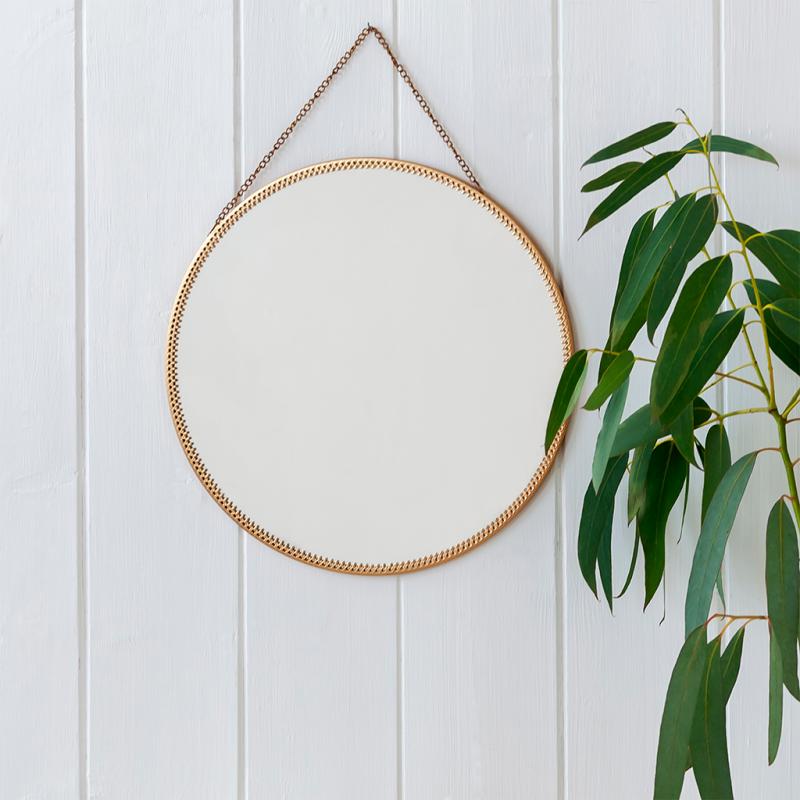 Circular gold tone mirror hanging on white wooden panel wall with green plant beside.
