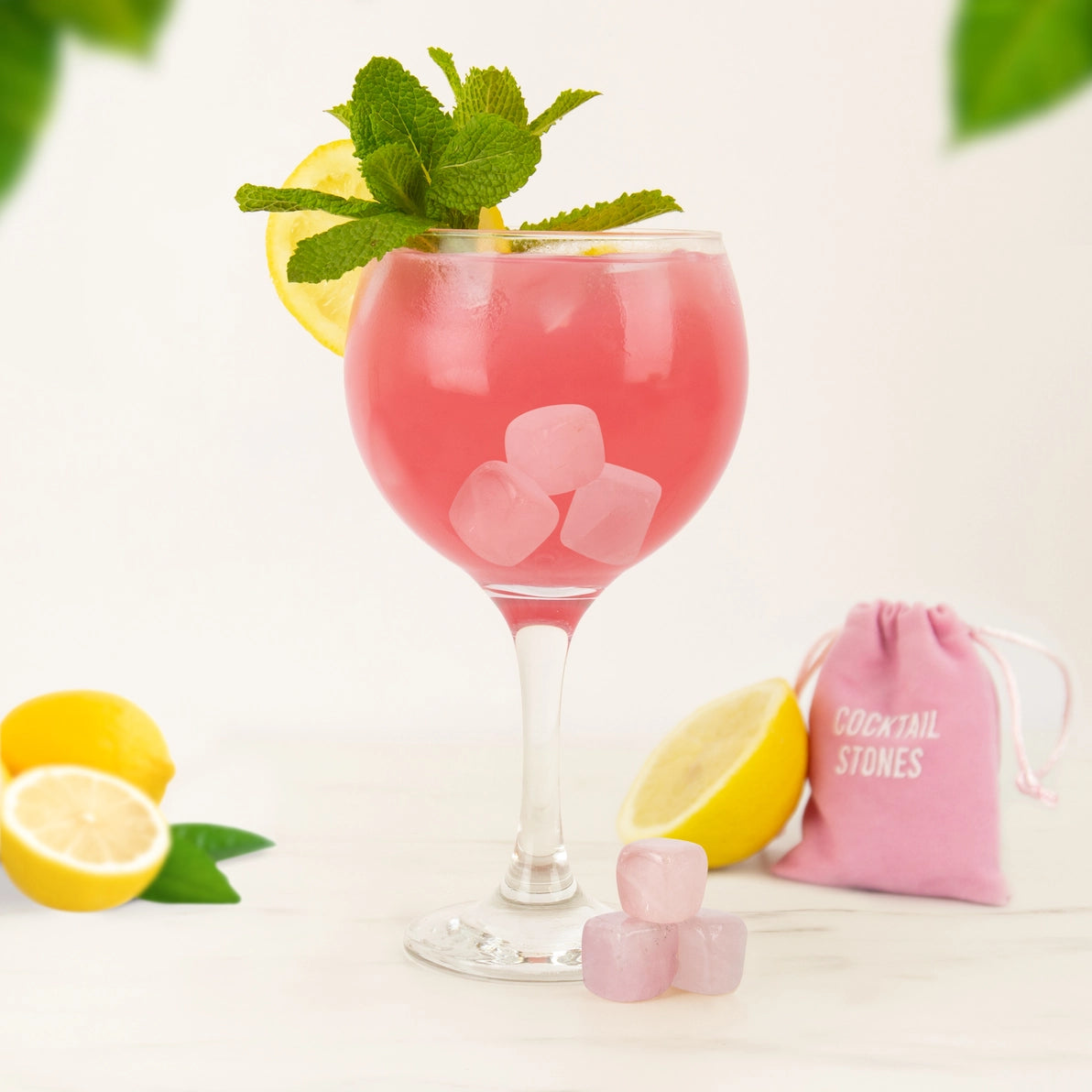 Rose quartz cocktail stones in a pink drink garnished with lemon and mint, beside a pink drawstring pouch and fresh lemons on a white surface.